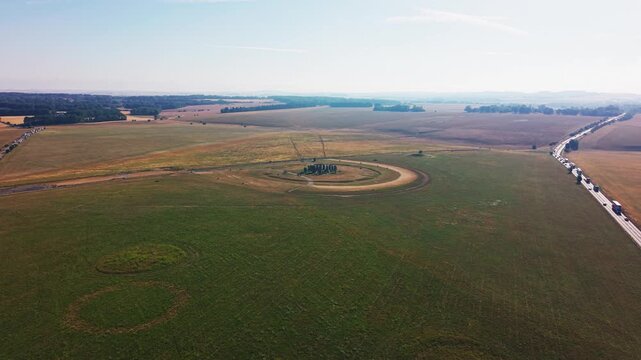 Wide aerial drone footage of Stonehenge surrounded by open fields on Salisbury Plain in Wiltshire, England. The elevated view shows the prehistoric stone circle, visitor traffic and a long queue