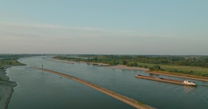 Drone orbits behind a cargo ship sailing along a Dutch canal, revealing dikes and expansive countryside farmland, showcasing inland shipping in a peaceful rural landscape.