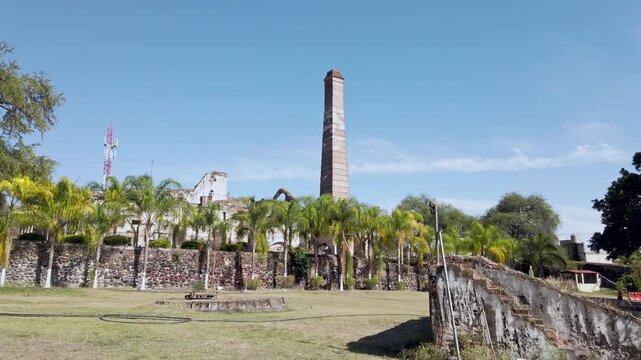 Ground view of the historic chimney at Hacienda Coahuixtla surrounded by palm trees, stone walls and preserved ruins under a clear blue sky in Morelos, Mexico