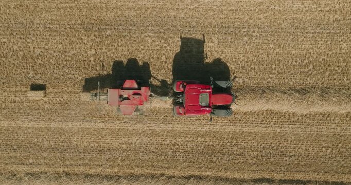 Top-down drone tracks a red tractor towing a straw baler, producing rectangular straw bales across a harvested wheat field in a clean geometric agricultural landscape and cinematic tractor shadow.