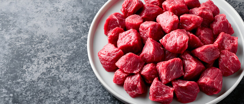 Top view flat lay of fresh raw diced beef meat cubes served on white plate over dark grey concrete background cooking recipe concept