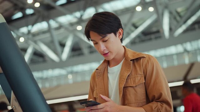 Young Asian man using smartphone at self service check in kiosk inside modern airport terminal, digital travel and passenger technology