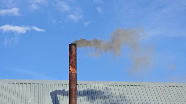 Thick black smoke from an old rusty chimney against a blue sky. Dark smoke rises from an old metal pipe above the roof of a building. Heating a rural bathhouse.