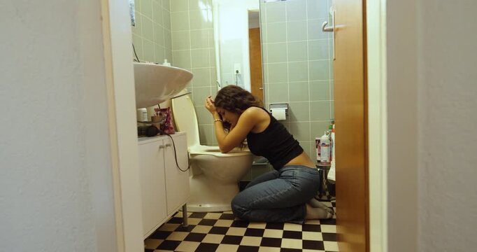 A woman kneels beside a toilet while feeling nauseous and unwell in a bathroom, suggesting symptoms of illness or stomach discomfort.