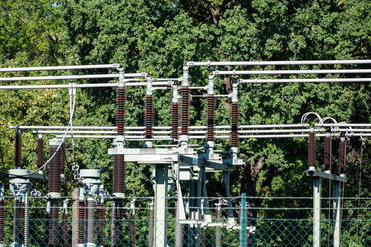 Detailed view of an electrical substation with complex power lines, high-voltage insulators, and metal structures in front of green trees. substation, power line, electricity, energy, infrastructure