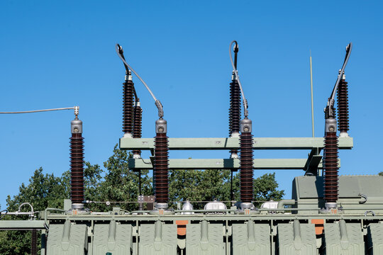 Detailed view of a high voltage transformer at an electrical substation under a clear blue sky, showing ceramic insulators, power lines, and green industrial infrastructure.