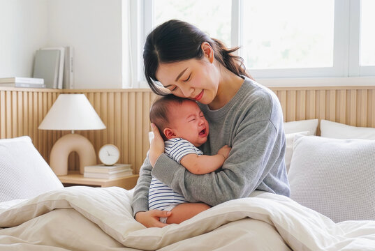 Caring Asian mother gently hugging and comforting her crying infant baby on bed. Parenting and maternal care concept