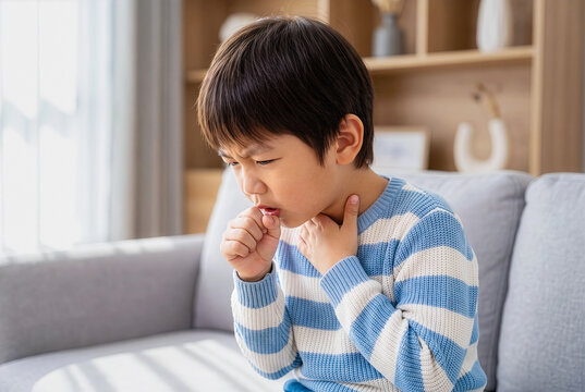 Sick Asian boy coughing and holding his throat, sitting on a sofa, suffering from a sore throat, cold, or flu symptoms