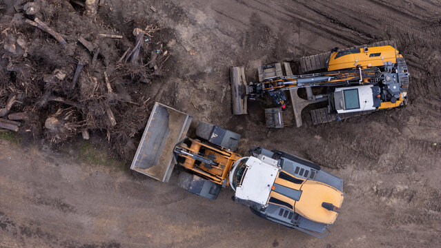 Aerial view of construction machinery including an excavator and a wheel loader clearing a large pile of tree stumps and roots on a dirt construction site in Gdansk, Poland.