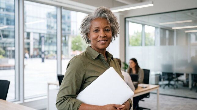 Senior adult woman holding laptop in modern office smiling at camera professional workplace diversity and inclusion