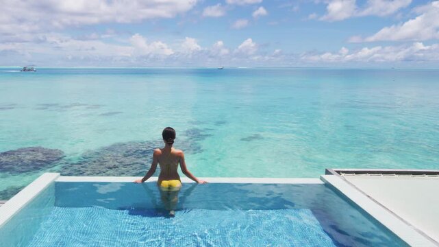 Travel vacation luxury resort idyllic overwater bungalow villa woman relaxing by infinity pool. Elegant beautiful lady in yellow bikini, by perfect blue ocean water
