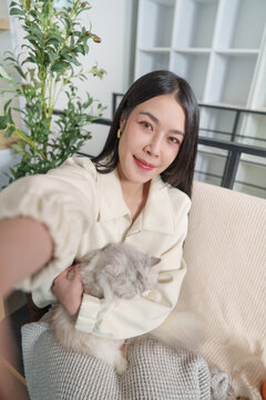 Asian woman taking selfie with fluffy cat on living room sofa