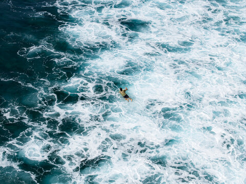 Aerial view of a surfer paddling on a surfboard amidst the turbulent white foam and turquoise waves of the ocean at Nusa Dua Beach in Kecamatan Kuta Selatan, Bali, Indonesia.