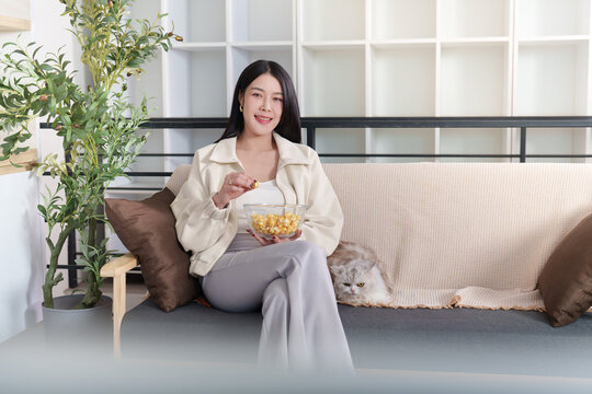 Asian woman eating popcorn from bowl while relaxing with cat on sofa