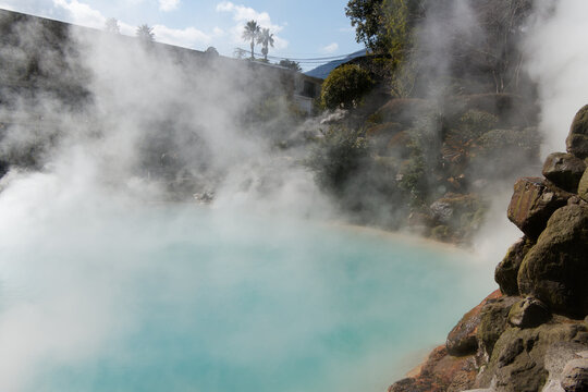 Thick white steam rising from a vivid cobalt blue geothermal hot spring pool surrounded by natural rocks and foliage.