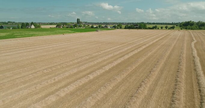 Drone flies above harvested wheat windrows while a red tractor bales straw across a Dutch countryside farm under a clear blue summer sky, showcasing modern agriculture and rural landscapes.