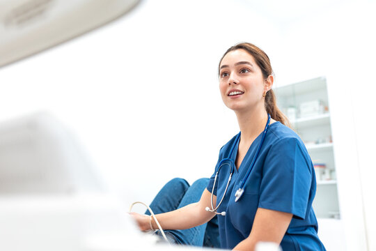 Female doctor sitting next to an examination table and operating an ultrasound machine in a bright clinic. Healthcare and medical diagnostic imaging examination technology concept.