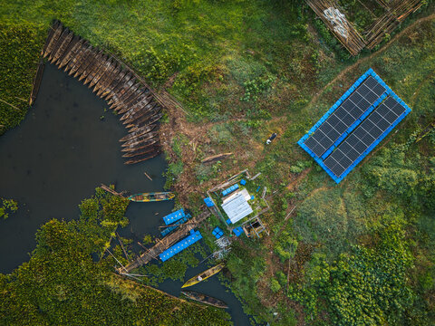 Aerial view of Lake Rawa Pening featuring a floating solar panel array, traditional wooden boats moored along the shore, and lush vegetation in Waduk Rawapening, Central Java, Indonesia.