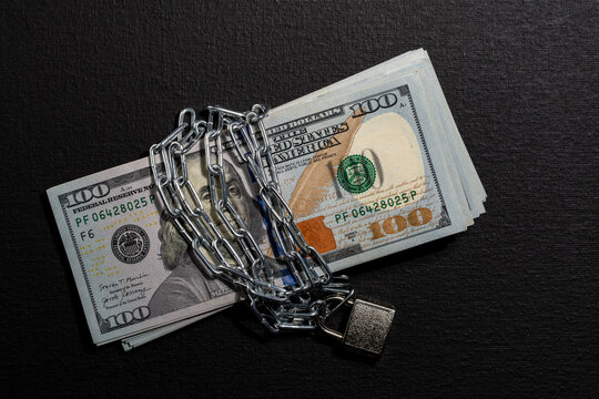US dollar banknotes wrapped in chain with padlock, closeup. Stack of hundred dollar bills secured with metal chain and locked padlock on black background