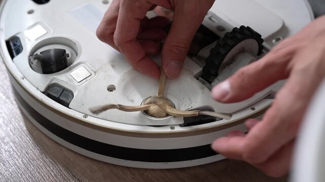 Detail cropped shot of male hands unscrewing and removing dirty, worn out side brush from robotic vacuum cleaner for replacement, highlighting home appliance maintenance and repair.