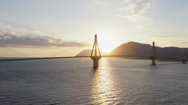 Rio-Antirrio Bridge, Greece. Drone pan at sunset. Bridge dramatically silhouetted. Sun hides behind pylons, backlit cinematic effect