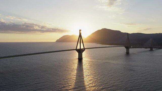 Panoramic drone flies toward Rio-Antirrio Bridge at sunset. Bridge silhouetted against glowing sky. Sun hidden behind pylon. Calm sea below
