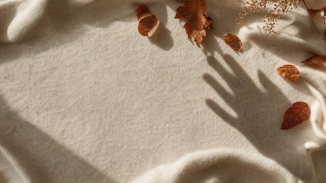 Dry leaves on beige wool fabric with human hand shadow. Cozy autumn background with soft light and texture. Minimalist seasonal flat lay for social media content or fall holiday.