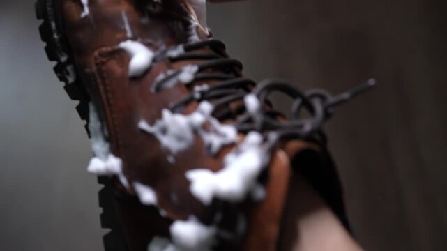 Hand using sponge to wash and clean dirty brown leather boot with soap foam, illustrating concept of footwear care and household chores. Concept of shoe care and maintenance practices, slow motion.