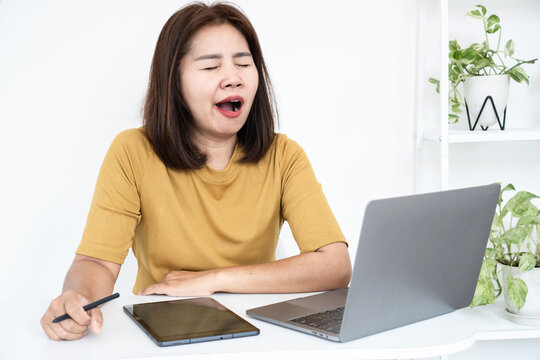 Exhausted tired Asian woman yawning at home desk while working on laptop computer, feeling sleepy, bored, lazy, fatigued, and burned out during work from home