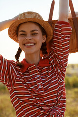 Obraz premium A smiling young woman in a striped shirt and straw hat carries a picnic basket outdoors. Lifestyle concept for summer leisure, joy, relaxation, and nature travel.