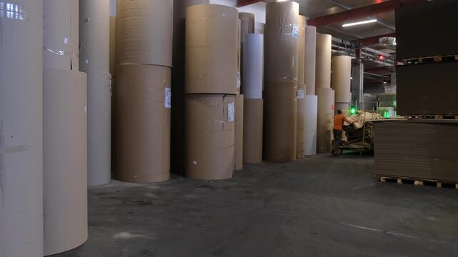A factory worker in protective wear pushes a metal cart loaded with scrap paper inside a spacious storage facility with massive paper reels.