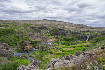 Fototapeta premium Scenic landscape of the lush green Gjain valley in Thjorsardalur, Iceland, featuring the Rauda river, small waterfalls, and volcanic rock formations.