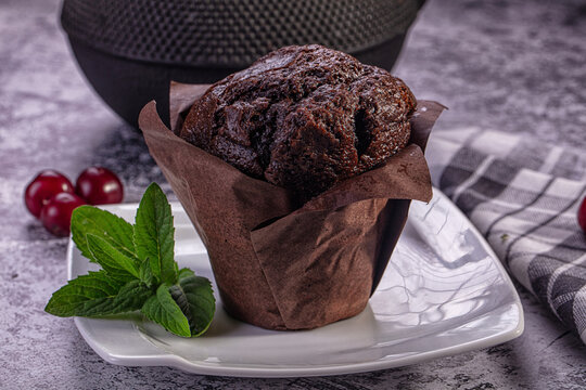 Chocolate muffin in brown paper liner on white plate with mint leaves and red berries