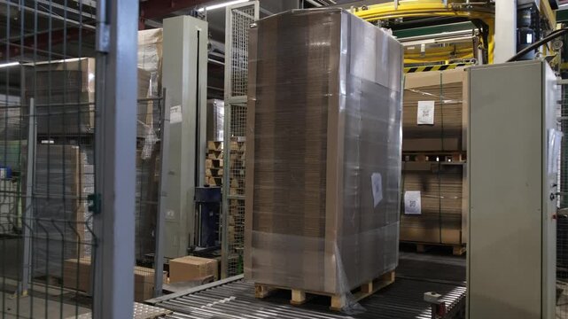 Industrial automated roller conveyor transport system moving a high stack of plastic wrapped cardboard sheets within a production facility.