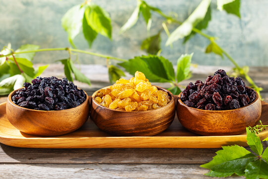 Close up raisin assortment with golden, black and dark red raisins in wooden bowls on rustic table with grape leaves