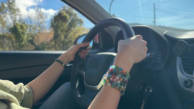 Woman driving car, hands on steering wheel with colorful bead bracelets. Static interior shot from passenger perspective looking forward.