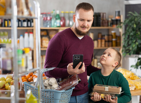 Attentive guy holding mobile phone and his son are looking for needed products in supermarket