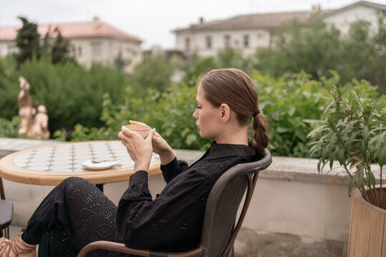 Pensive woman holding coffee cup while sitting on outdoor terrace overlooking urban scenery