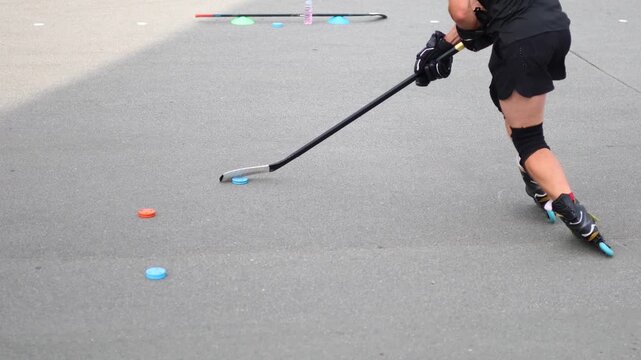 Slow motion cropped 4k video: legs of a middle aged man practices hockey stick techniques while roller skating on an asphalt rink in the summer sunny weather.
