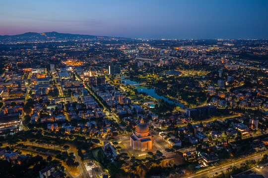 Aerial view of the illuminated Basilica dei Santi Pietro e Paolo in the EUR district at twilight, surrounded by urban architecture, city lights, and a park in Rome, Lazio, Italy.