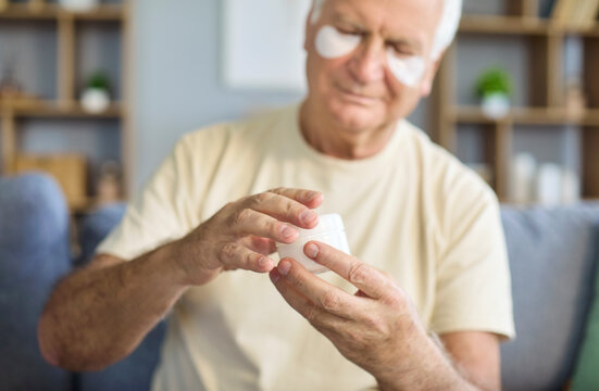 Senior man applies beauty cream with eye patches. At home he follows a grooming routine for hydrated face, easing puffiness and lines for healthy skin. Symbolizes senior skincare and wellness.