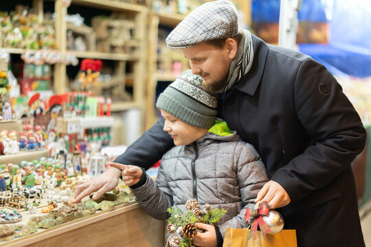 Dad and son are standing near counter at Christmas market fair, choosing figurines for nativity scene. Customers look at product with interest, select right figures. .