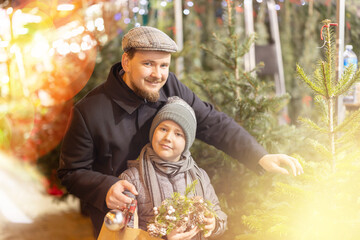 Obraz premium Dad and son are looking at natural and artificial Christmas trees at street fair. Family walks around bazaar, examines goods, and stares at festive assortment of fair. .