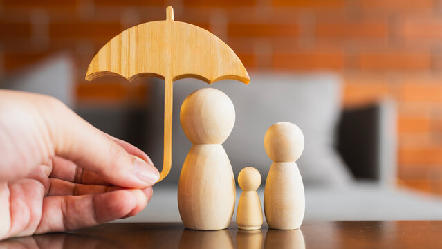 Hand holding small umbrella over wooden family figures representing insurance, home protection, and financial security.