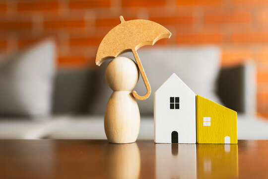 Wooden figure under umbrella with toy house models representing real estate insurance, mortgage protection, and loan.
