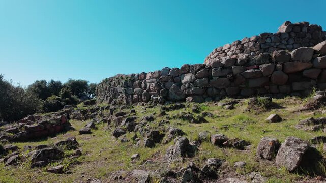 A wide shot of the prehistoric Nuraghe SOrtali e su Monti archaeological site in Sardinia, Italy, featuring ancient dry stone walls under a clear blue sky.