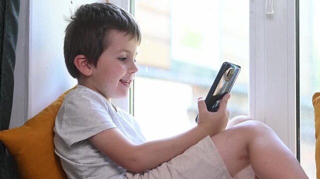 A 6-7 year old child sits on a windowsill at home with a mobile phone and plays. Mental health issues, unlimited gadgets, FOMO, harm to the child's psyche.