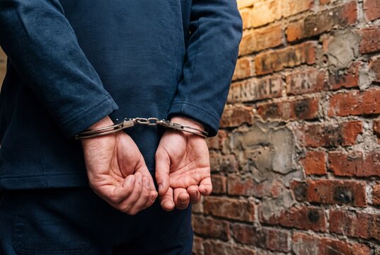 Close up of a man in steel handcuffs with hands restrained behind his back against a weathered brick wall in warm light with no face visible, representing arrest crime and criminal justice concept
