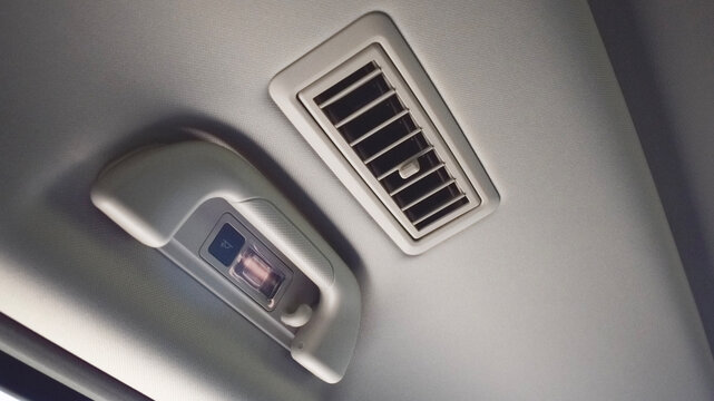 Close-up view of a modern car interior ceiling featuring an air conditioning ventilation grille vent alongside an overhead passenger grab handle with built-in reading light and coat hook