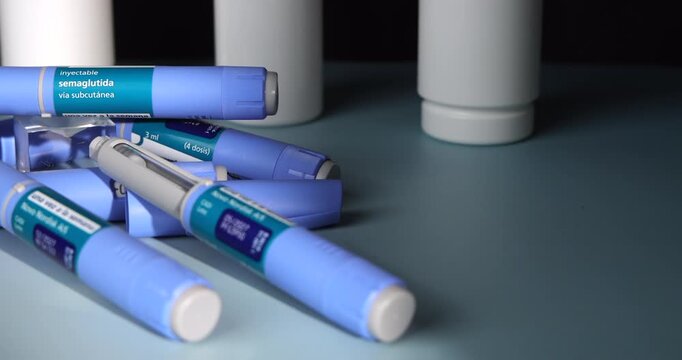Slow horizontal camera dolly movement sweeping across blue GLP-1 Semaglutide pens alongside white prescription pill bottles on a light blue table surface with a dark black backdrop, medical view